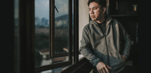 an asian chinese mid adult man looking outside of window from his home during sunset with serious facial expression