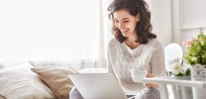Smiling women in a light filled living room drinking tea and looking at her laptop.