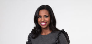 A smiling woman with shoulder-length dark hair, wearing a grey top with ruffled sleeves, poses in front of a plain light-colored background