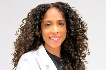 Jasmine A person with long curly hair wearing a white lab coat and black shirt smiles at the camera against a plain background